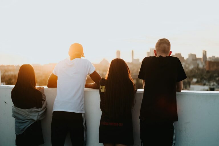 Groupe de jeunes de dos, accoudés à un muret face à la ville au coucher du soleil