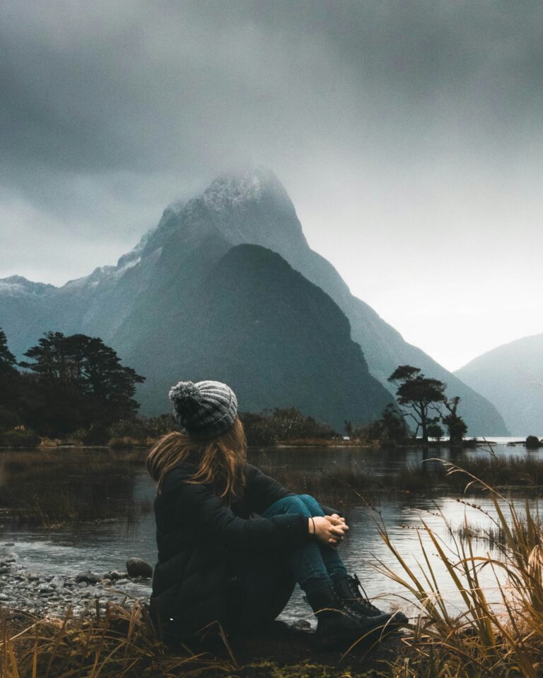 Jeune femme assise au bord d’un lac, face à une montagne sous un ciel gris